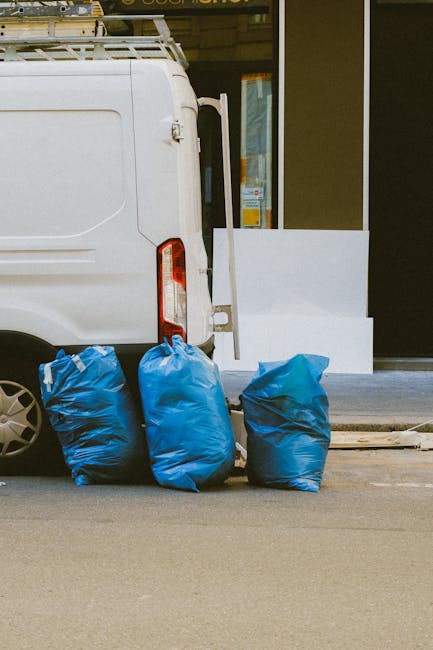 A white commercial van parked on a paved urban street with its rear door closed, positioned next to three large blue rubbish bags filled with waste material placed directly on the ground. The bags are made of durable plastic with a slightly crinkled texture, and they vary in shading from light to darker blue. In the background, part of a modern building features a dark, smooth facade with a glass window or door frame, and a white panel or barrier is situated near the entrance. The scene is lit by natural daylight, providing clear visibility of the objects, and the setting suggests a typical environment where private waste collection or on-site rubbish disposal might be arranged outside a commercial or residential premises. Waste Removal Ealing appears to be involved in handling the rubbish indicated by the cohesive arrangement of the waste bags and van, aligning with alternative approaches to rubbish collection in support of efficient waste management and disposal practices.