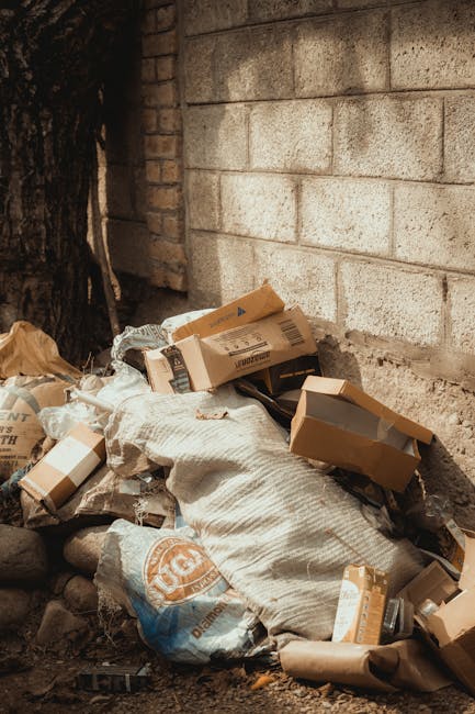 A pile of mixed waste, including cardboard boxes, paper packaging, and plastic bags, is gathered against a concrete brick wall outdoors. The cardboard boxes vary in size, some partially flattened, with visible printed labels and brown kraft finishes. The paper packaging appears crumpled and is partly covered by a white sack made of woven material. Surrounding the waste are small rocks and dirt, indicative of an exterior setting such as a corner of a building or a back alley. To the left, part of a tree trunk is visible, contributing to an outdoor environment. The scene is illuminated by natural light, casting soft shadows across the debris, illustrating an area prepared for private rubbish collection or on-site waste clearance, typical of services offered by waste removal companies like Waste Removal Ealing.