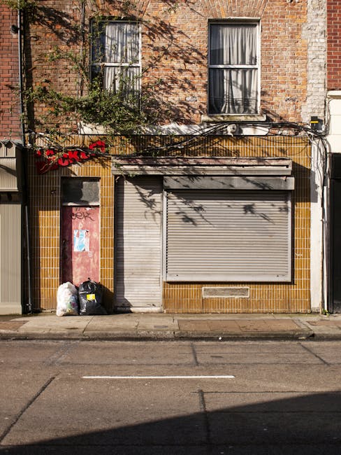 The image depicts the front exterior of a building with a brick facade and two vertically aligned windows with curtains on the upper floor. Below, the ground-level storefront features a closed, grey roller shutter and a narrow door on the left, both set into a wall covered with yellow-brown ceramic tiles. To the left of the door, there are three black garbage bags placed on the pavement, alongside a white plastic bag and some smaller debris, indicating waste awaiting collection. Above the storefront, tangled black cables run horizontally across the building, partially casting shadows on the brick surface. The pavement is unmarked, with a slight shadow at the bottom of the image, and the scene suggests an urban environment where waste collection or private rubbish removal services might be needed, as indicated by Waste Removal Ealing’s area of operation. The overall scene appears quiet and clean aside from the waste bags, highlighting the importance of effective rubbish disposal in such settings.