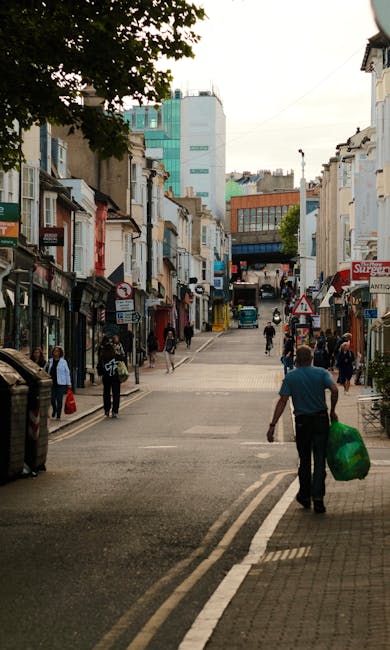 A wide city street scene in an urban area with a mix of modern and older buildings on both sides. The foreground shows a man walking away from the camera, carrying a green bag of waste, indicating a rubbish removal or disposal activity. The street is lined with small retail shops with signage, some with awnings, and pedestrians are walking along the pavement on either side. There are parked cars and a few people entering or leaving shops, with a larger modern building visible further back, contrasting with the smaller storefronts. The environment appears busy but orderly, with clear weather and daylight illuminating the scene. The overall setting suggests a typical urban thoroughfare where residents and businesses might seek private rubbish collection services, such as those offered by Waste Removal Ealing, to handle waste disposal needs beyond standard municipal collections.