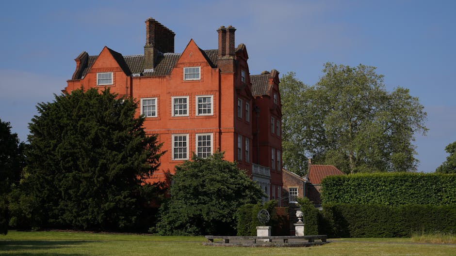 A large, red-brick Victorian-style building with multiple gabled roofs and numerous rectangular white-framed windows dominates the background. The building has a weathered finish typical of historic architecture and features several prominent chimney stacks extending from the roof. In the foreground, there is a well-maintained lawn with lush green grass, bordered by mature trees and dense shrubs, including a prominent large evergreen tree to the left and a deciduous tree on the right with fresh green foliage. A neatly trimmed hedge runs along the right side of the scene, creating a natural boundary. Near the base of the building, a stone or concrete staircase with decorative urns or planters on pedestals is visible, possibly part of a garden or terrace area. The overall setting appears to be a landscaped garden or park area, with soft daylight illuminating the scene, and the environment suggesting a quiet, residential, or institutional property. The scene subtly aligns with private waste management and rubbish removal services, which might be involved in maintaining such properties, emphasizing careful landscaping and waste clearance efforts.