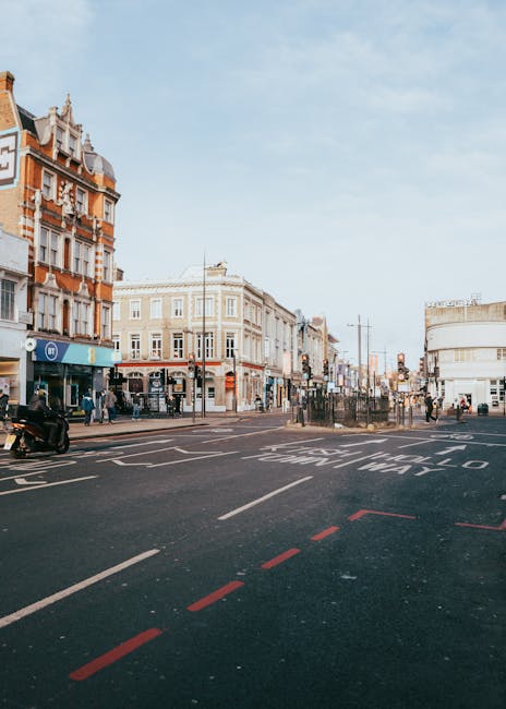 A wide view of a busy urban street scene featuring a row of multi-storey buildings with ornate architectural details and a mix of brick and cream-colored facades on the left side. The buildings house various shops, including a visible blue retail outlet at the corner, and pedestrians are walking along the pavement. In the foreground, a section of asphalt road is marked with white lane lines and red dashed lines, indicating parking restrictions or designated areas. A motorcycle is parked near the curb on the left, and a few pedestrians cross or wait at the crosswalk further along the street. The background shows more commercial buildings, street furniture, and clear sky, suggesting a bright, daytime environment. This scene is characteristic of a central shopping district that might be an example of an area where independent waste collection services, such as those by Waste Removal Ealing, could facilitate on-site rubbish clearance for local shops and businesses, providing an alternative to traditional municipal waste services.