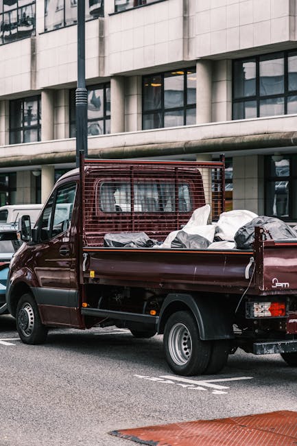 A small, dark red flatbed truck parked on a city street in front of a modern building with large glass windows and a concrete facade. The truck's cargo bed is filled with various types of waste, including several large white and black plastic bags, some appearing to contain household rubbish or miscellaneous debris. The bed has a metal frame at the front with a grid pattern, likely to prevent load spillage. The vehicle's tires are black with standard silver wheel rims, and the rear taillights and license plate are clearly visible. Surrounding the truck are other parked vehicles, including a white van, with the scene illuminated by natural daylight, conveying an environment suitable for private waste collection or on-site clearance activities provided by Waste Removal Ealing, especially as an alternative to traditional rubbish disposal methods.